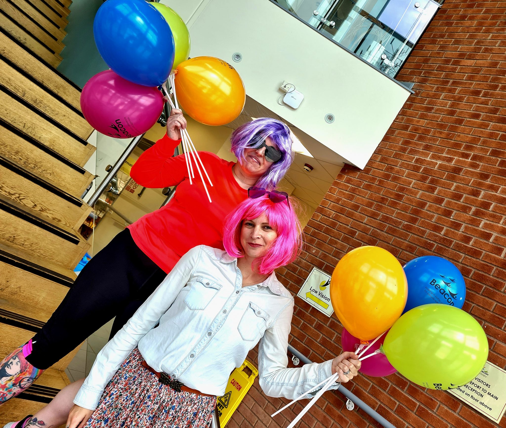 This image shows Mary and Sarah from Beacon stood in the reception area of our centre wearing brightly coloured clothes, wigs and balloons.