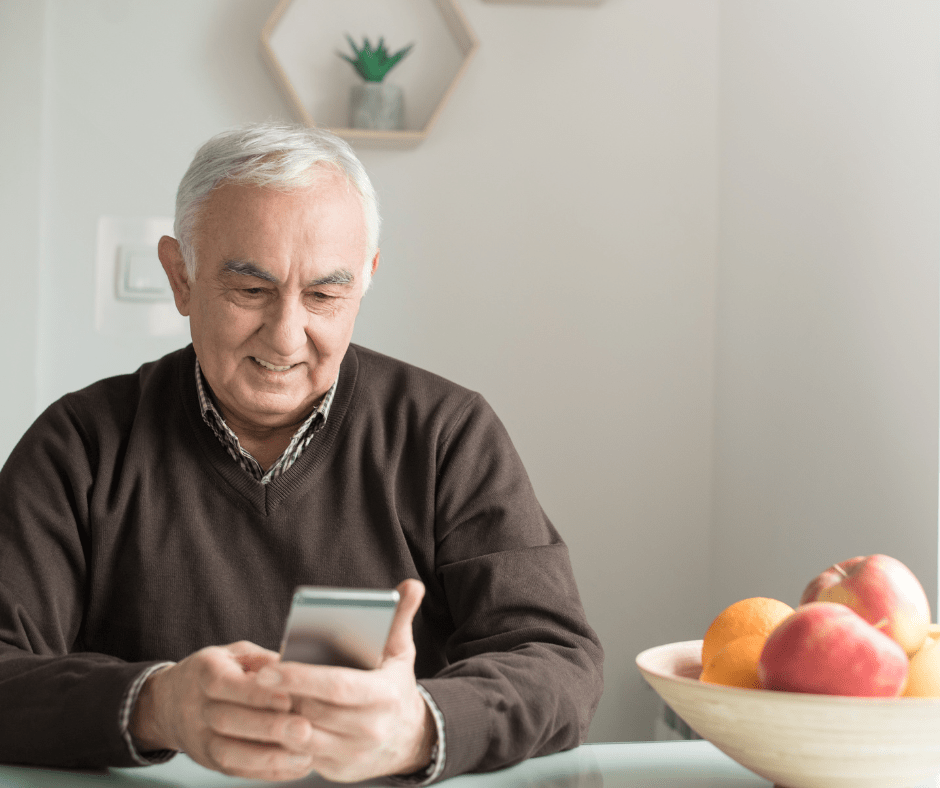 This shows an older man sat at a kitchen table looking at his mobile phone and smiling,