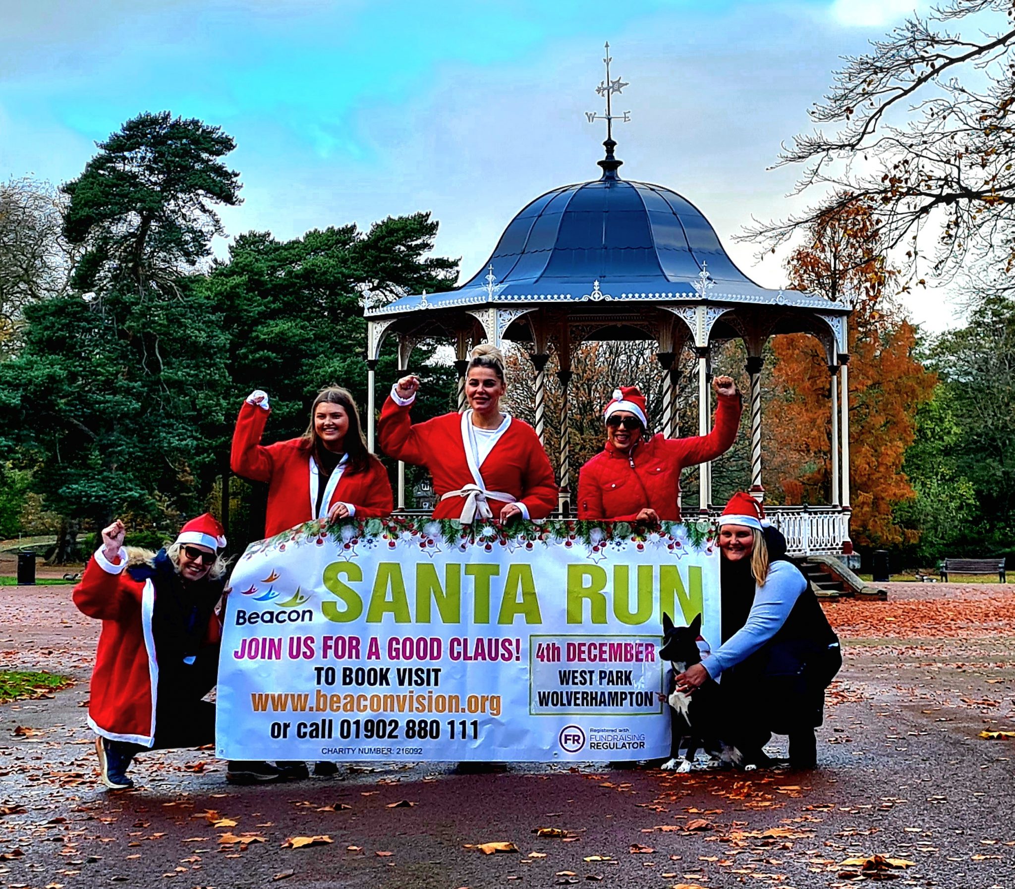 There are five women on this picture and a dog. They are all dressed in Santa outfits and they are holding a large banner promoting our Santa Run. They are in West Park and behind them is a bandstand.