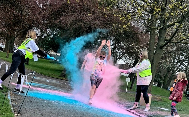 This shows a participant in our Colour Run walking through a colour station on the route as blue and pink powder is thrown on him.