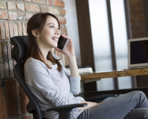 A lady holding a mobile phone to her ear, sat in an office chair wearing a grey top and jeans.