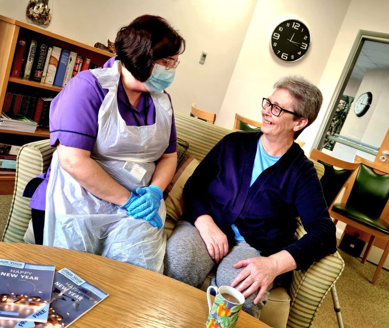 A member of our care team dressed in purple uniform and PPE, talking with a member sat on green sofas in coffee lounge.