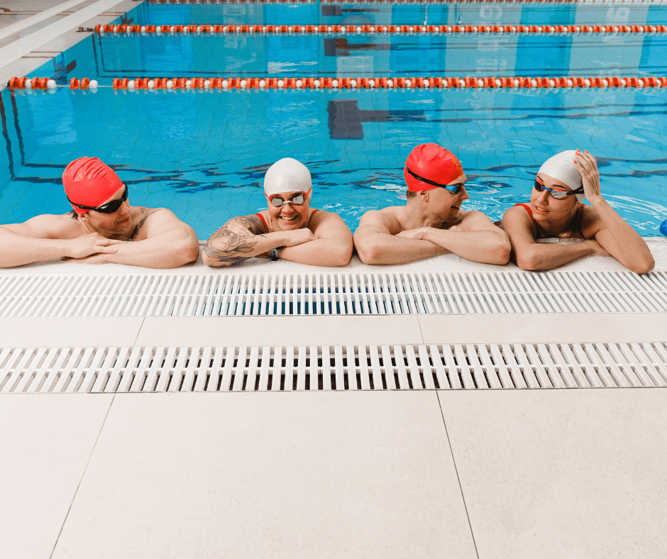 This shows four adults in swim hats and goggles leaning against the side of a pool.