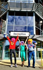 This shows Jarad, Sarah and Steve from Beacon stood outside our centre reception. They are all wearing brightly coloured wigs and have floral garlands around their necks.