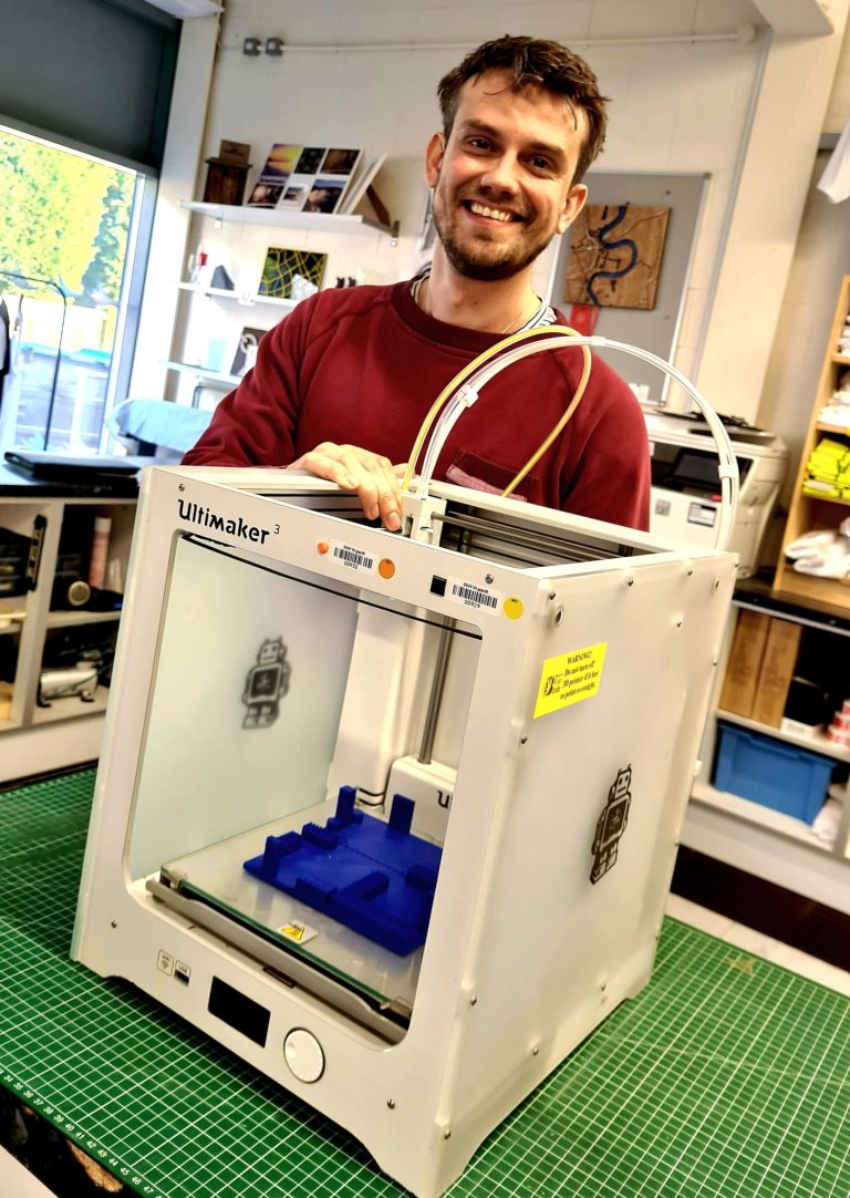 A man is smiling wearing a red jumper standing behind a 3D printer in the Fab Lab