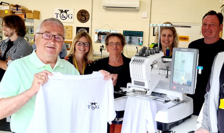 A group of people in Beacon's Fab Lab standing behind a new embroidery machine, with one holding up a white t-shirt.