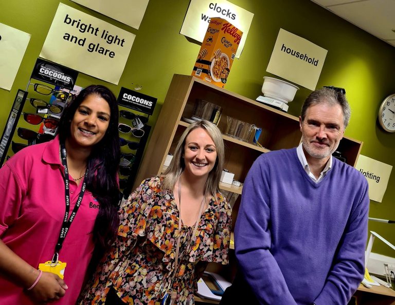 Three staff members in front of assistive tech equipment smiling at the camera