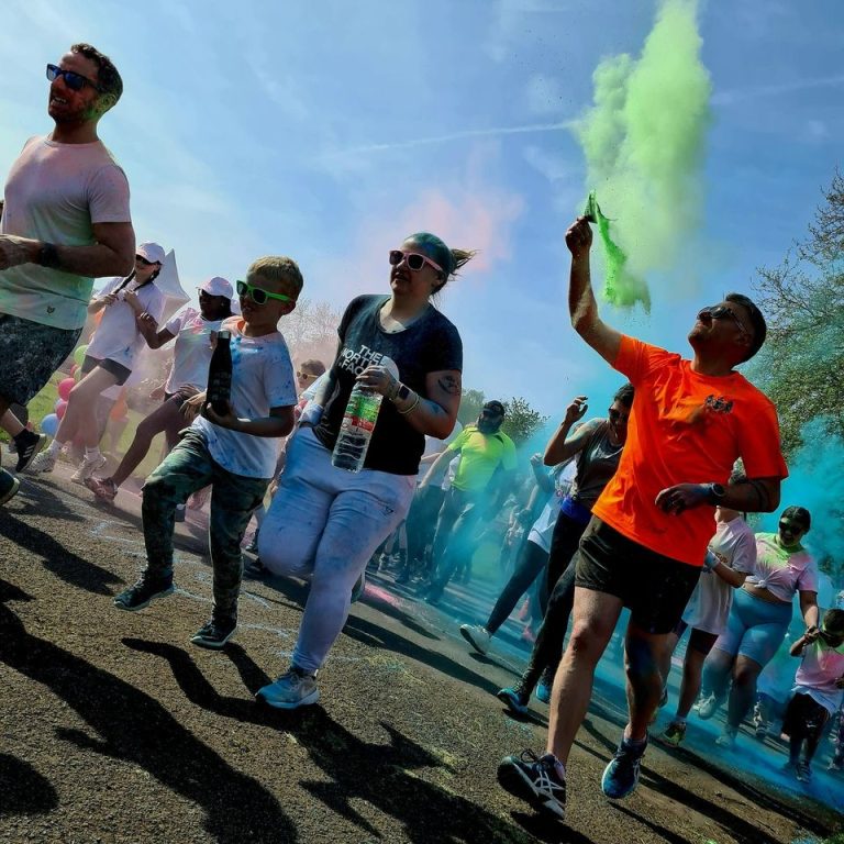 This shows a shot of participants at our Colour run setting off. There is a man with an orange t-shirt who is letting off green powder into the sky.