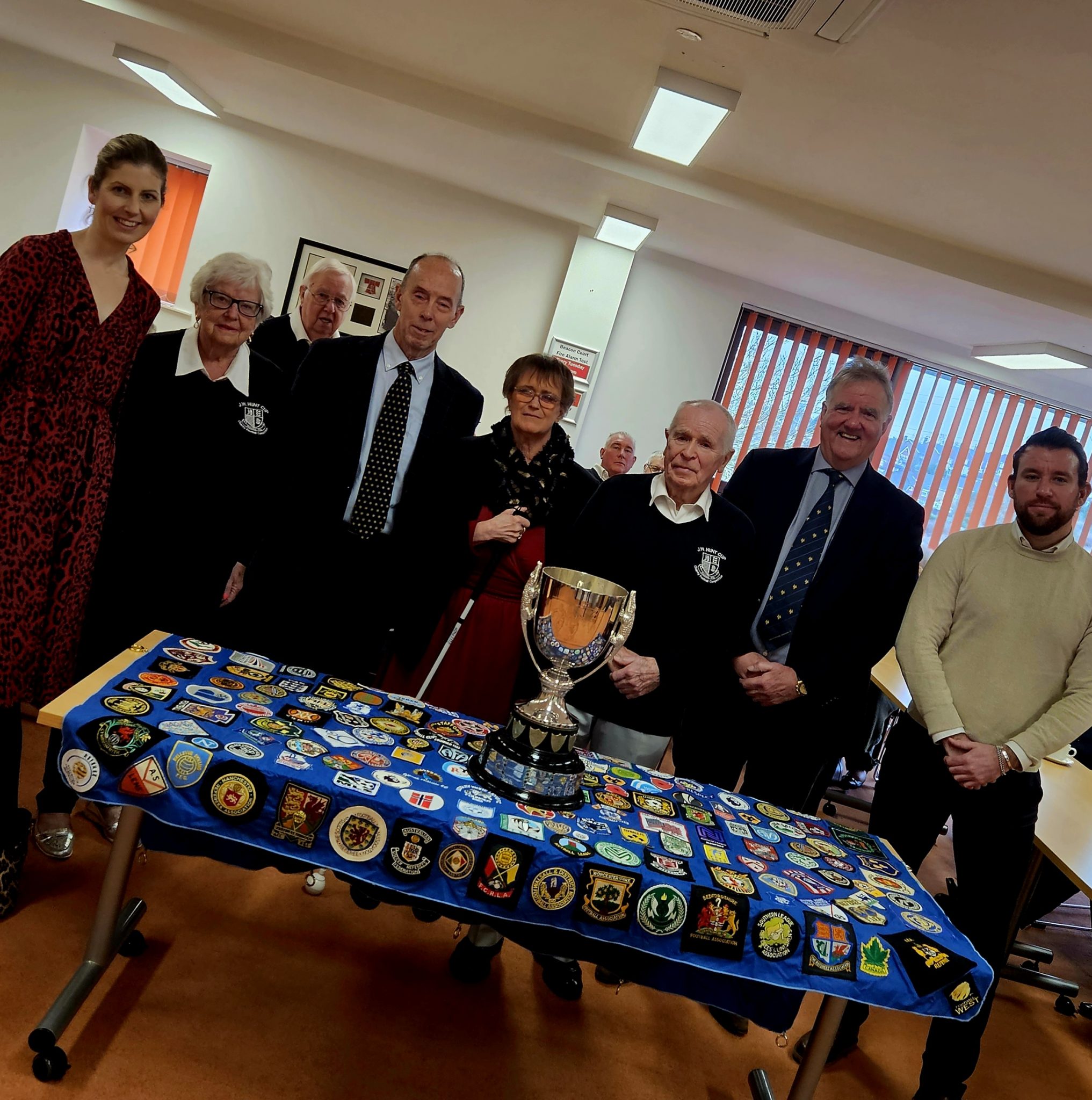 This picture shows the cup on a table covered with the club crests of teams which have taken part in the cup. Behind it are members of the JW Hunt Cup committee, Gough Group Holdings and representatives from the Beacon Centre.
