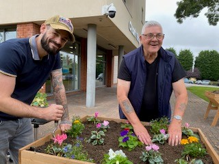 This shows two men, standing over a planter outside our centre planting shrubs and flowers, They are both looking towards the camera, laughing.