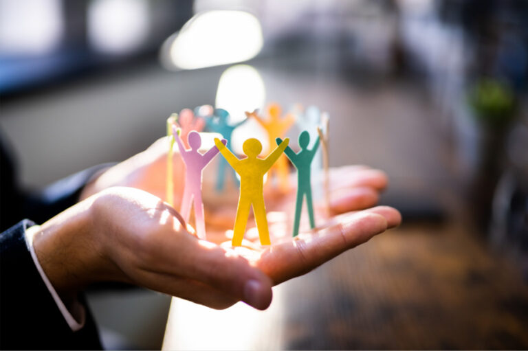 This shows two cupped hands holding a circle of cut out brightly coloured people. The background is blurred.