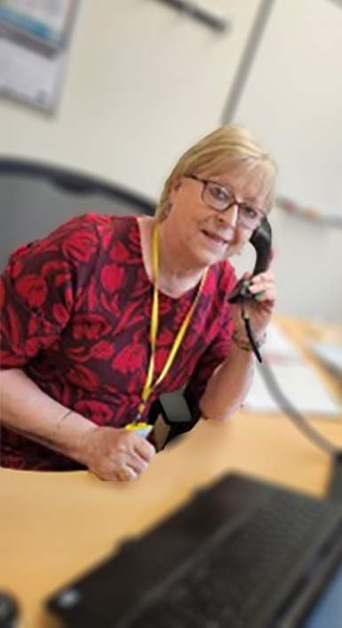 This shows Judy from Beacon, she has short blond hair and wearing glasses and is wearing a red dress. She is speaking on the phone as she smiles to the camera.