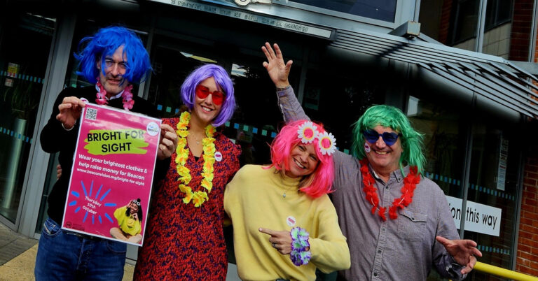 This shows four people, two men and two women dressed in bright clothing and wigs standing outside our centre. A man on the end is holding a promotional poster.