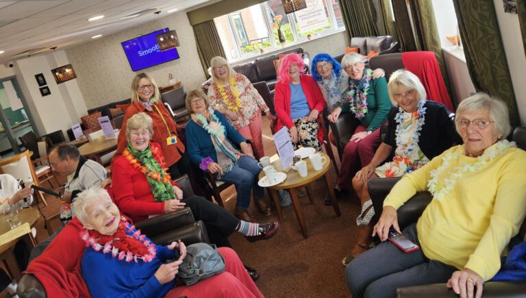 This is a group of women from the Sedgley Morning Townswomen Guild dressed up in brightly coloured outfits sat in a coffee bar.