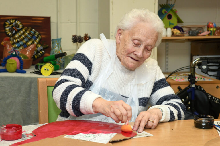 This shows one of our members in our craft room painting a cardboard guitar brown. She's using a sponge paint brush and is smiling.