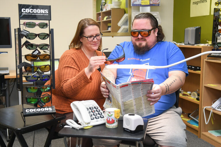 This shows a man getting advice about lighting and glasses at our centre by one of our Sight Loss Advisors. in front of them are two tables with items including Cocoon glasses, a phone with large buttons, a talking clock and a mug with a liquid level indicator. He's reading a tv listing magazine under a bright light.