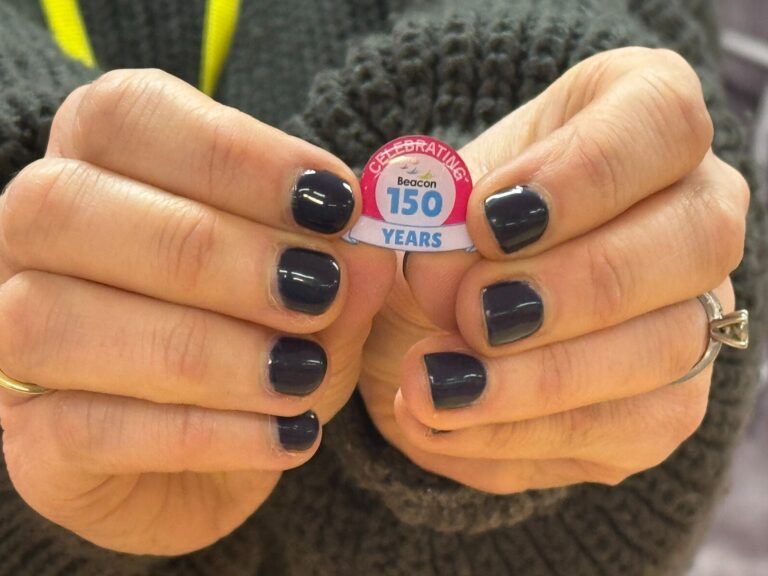 A pair of hands with dark nail polish holds up a round pin with the words "Celebrating Beacon 150 Years" written on it using the winning design from the internal competition