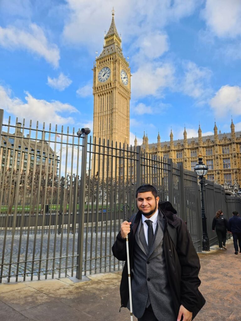 Portrait of Muhammed smiling on a London pavement near Big Ben, dressed formally and holding a white mobility cane, with the Palace of Westminster behind him.