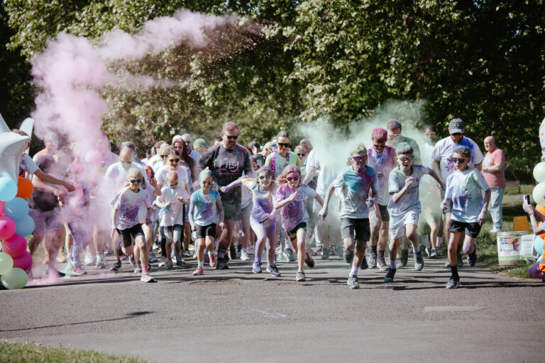 Children and adults run forward at the start of a colour run on a park path, surrounded by clouds of pink and green powder, with spectators and trees in the background.